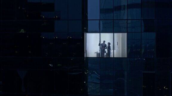 从摩天大楼外的航拍镜头:商人和女商人站在办公室窗户里交谈找回傍晚商业区的飞行镜头和在大城市工作的商人