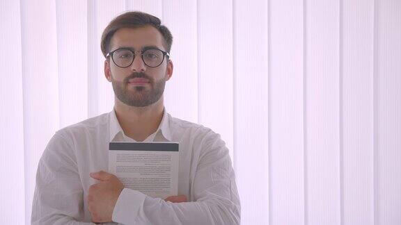 特写英俊的成功的白人商人戴着眼镜拿着一本书看着相机微笑在室内白色的办公室