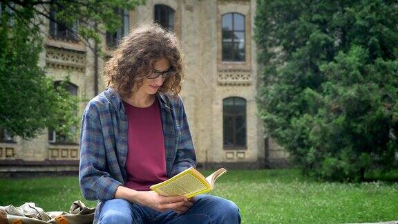 戴眼镜、卷发的帅哥坐在大学附近公园的长椅上看书认真而专注
