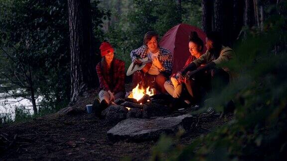 一名男性游客正在弹吉他而他的朋友们则在森林里的帐篷附近一起坐着一边听一边往篝火里扔柴火音乐和友谊