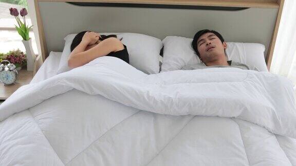 亚洲年轻的妻子不能睡觉她的丈夫在她旁边打鼾妇女关闭耳朵与枕头