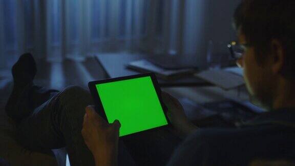 男人晚上躺在沙发上在风景模式下使用绿色屏幕的平板电脑