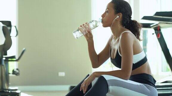 迷人的年轻运动女性在室内健身房里的跑步机上跑步喝水休息跑步女孩穿着运动服听着音乐坐在跑步机上锻炼