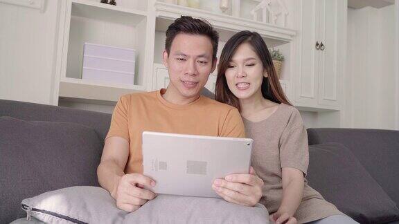 亚洲情侣在家中客厅用平板电脑上网购物甜蜜情侣在家中放松时躺在沙发上享受爱情时刻生活方式夫妇在家里放松