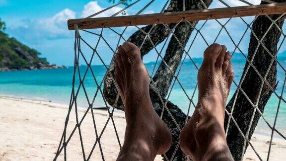 在白色的沙滩和蓝色的海洋前赤裸的男人的脚在吊床上晃荡
