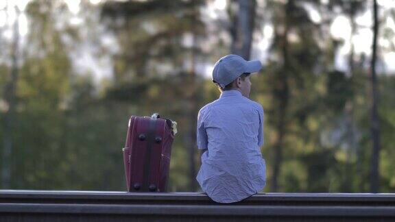 戴着帽子的伤心男孩坐在外面铁路上的一个手提箱上