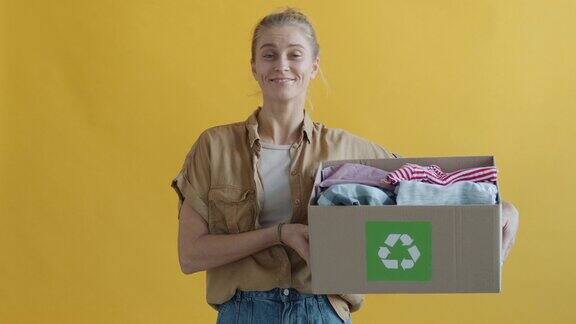 一名年轻女子拿着装着旧衣服的纸板箱在黄色背景上进行回收