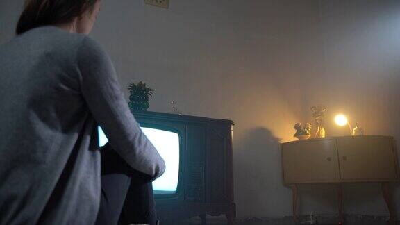 年轻女孩坐在老房间的地板上看复古电视的背影复古家具装饰暗室老式电视机打开翻动复古风格的电视屏幕家中破旧的老式设备复古的电视