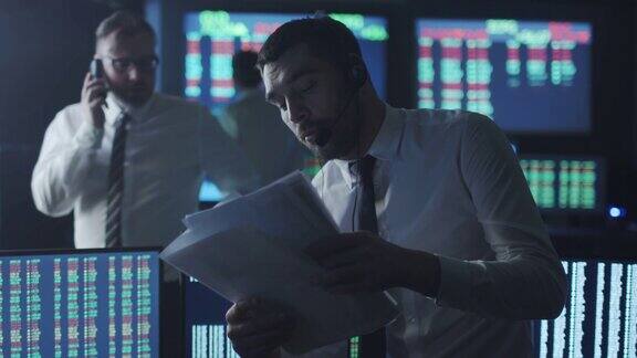 一名身穿白衬衫的股票经纪人正在一间有显示屏的黑暗监控室里打电话