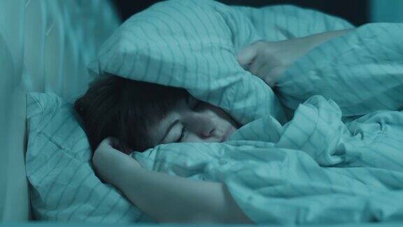深色头发的女人被噪音干扰难以入睡用枕头捂着耳朵感觉压力很大试图入睡饱受失眠之苦