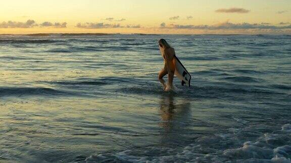 穿着泳衣的美丽年轻女子背着冲浪板走进大海她看起来美丽和苗条有一头金发大海浪和日落的背景