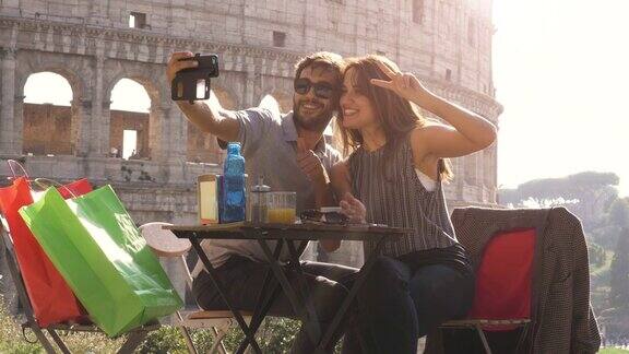 一对快乐的年轻游客夫妇在日落时分用智能手机自拍坐在罗马斗牛场前的酒吧餐厅里带着咖啡购物袋笑着玩
