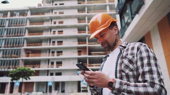戴着防护头盔的白人男性建筑工人在正在建造的建筑附近使用智能手机工程师检查施工过程在电话上做记录领班聊天应用
