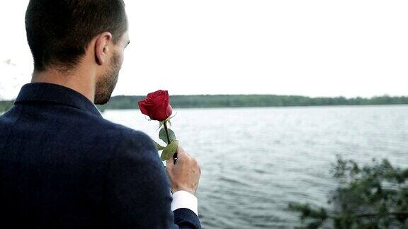 一个穿着西装的年轻商人在湖边手里拿着一朵红玫瑰