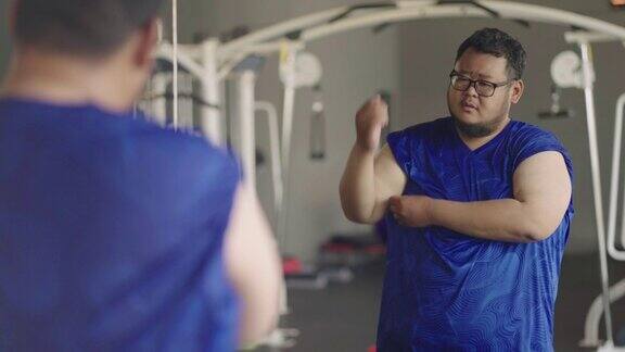 在室内健身房一个非常胖的男人站在镜子前看着自己拉起他的衬衫摇晃着他的啤酒肚和前臂