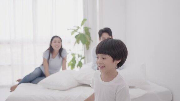 亚洲幸福家庭爸爸妈妈和可爱的儿子在舒适的家里享受周末爸爸和妈妈一起坐在舒适的床上微笑着看着儿子抱着枕头跳来跳去玩得很开心慢动作镜头