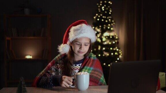 快乐的女孩戴着喜庆的红色圣诞帽穿着格子呢坐在笔记本电脑上看圣诞电影房间内部装饰得很漂亮新年假期和儿童概念