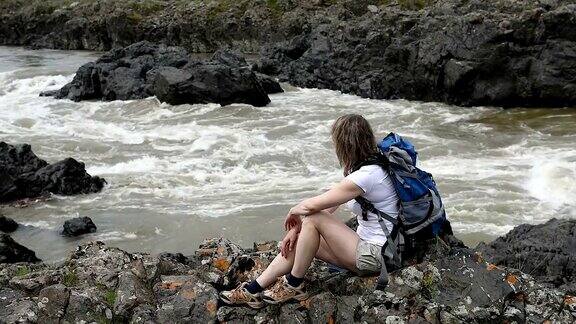 一个女孩背着背包坐在山河岸边的一块岩石上