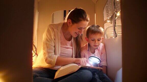 微笑的年轻母亲和小男孩读书在晚上玩玩具屋孩子的教育和家庭在晚上有时间在一起