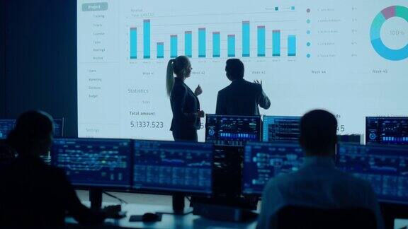 项目负责人、首席执行官讨论大屏幕显示的数据屏幕显示信息图表图表财务分析股票市场增长有工作人员的电信控制室