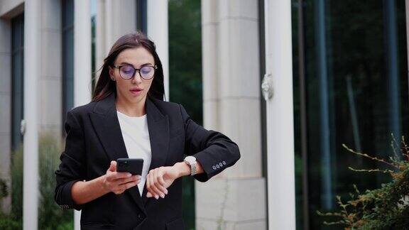 一位年轻的女商人赶着去参加一个会议在一座现代化的办公楼附近看了看表忙碌的女办公室职员穿着经典的西装戴着眼镜在户外上班迟到了