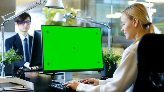 在办公室里女商人在她的办公桌前对着一台模拟的绿色屏幕的个人电脑工作同事坐在她对面现代看房间与玻璃和混凝土风格