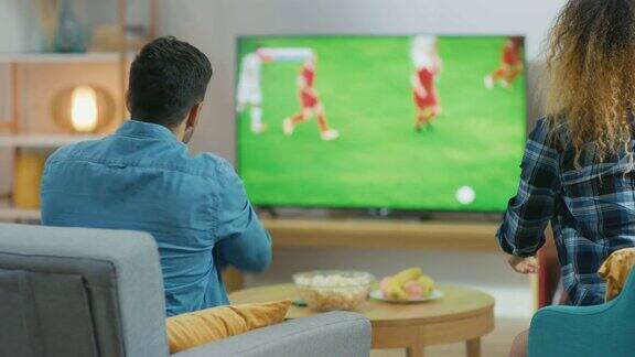 在家里一对年轻的夫妇在电视上观看足球比赛他们在情绪上担心和欢呼在进球得分后为他们的球队的胜利而击掌庆祝后视图相机拍摄