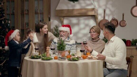 家庭年夜饭餐桌上一个戴圣诞帽的老人和一个戴眼镜的女人两个孩子一个男孩一个女孩两个年轻人男人和女人拿着酒杯女孩把盘子放在桌子上