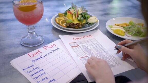 营养学家女子写饮食计划与食物卡路里表在桌子上与潘趣酒和水果盘在咖啡馆