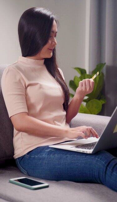 亚洲妇女坐在沙发上拿着信用卡使用笔记本电脑在家里网上购物