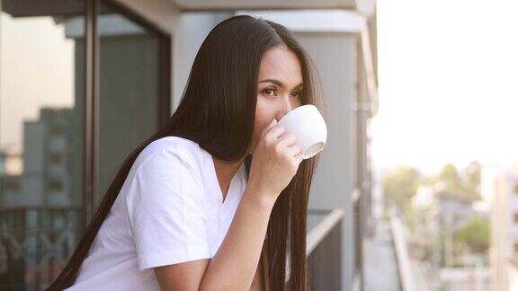 一个亚洲女人正站在阳台上喝着热咖啡在早上