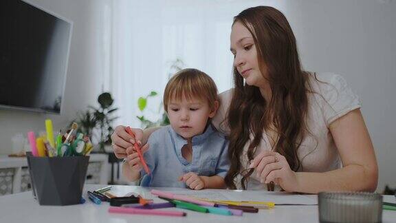 一个有两个孩子的家庭和一个年轻的母亲坐在桌子旁用彩色铅笔在纸上画画儿童创造力的发展白色干净的室内