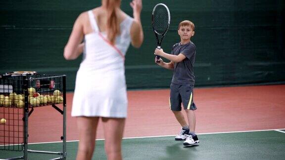 女教练教小男孩打网球穿着白色运动服的妇女站在球场上向孩子扔黄色的球年轻的运动员正在学习用球拍回网球