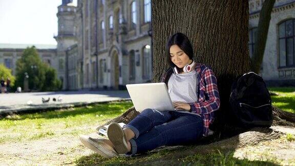 年轻的大学生使用笔记本电脑坐在树下微笑社交