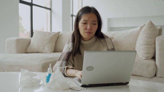 一个亚洲女人用餐巾擤鼻涕坐在家里的办公桌旁用笔记本电脑学习一个生病的女人得了流感用毯子盖住自己得了感冒
