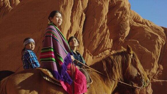 几个年轻的印第安人(纳瓦霍)孩子穿着传统的纳瓦霍服装和坐在他们的马看着亚利桑那犹他州纪念碑谷沙漠的景观旁边的一个大的岩石形成在一个缓慢的移动镜头
