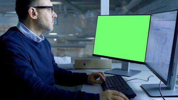首席男性工程师在他的个人电脑上工作的技术项目第二显示显示绿色屏幕模型从办公室的窗户可以看到大工厂