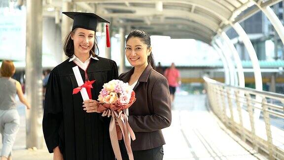 美丽的微笑毕业的学生女孩的肖像年轻的女人和她的母亲在帽子长袍走和拿着文凭