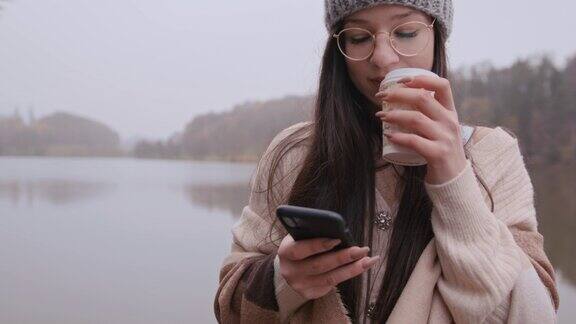 年轻女子在湖边一边用纸杯喝咖啡一边用手机
