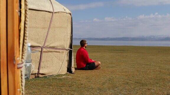在吉尔吉斯斯坦一个穿着红色衣服的旅行者坐在可以看到宋库尔湖和山脉的蒙古包营地迎接明媚的早晨
