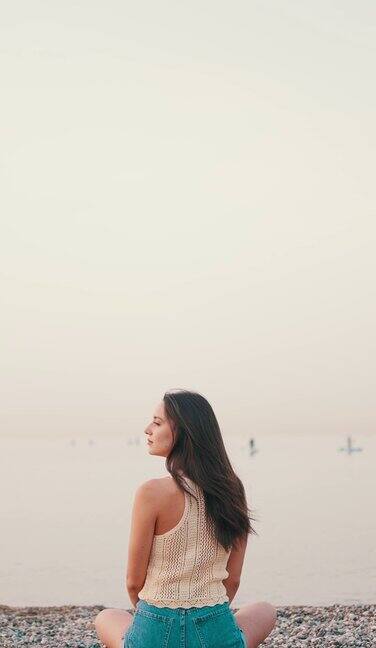 垂直视频:美丽的棕色头发的女人长头发坐在海滩上的海景背景华丽的女孩穿着上衣和牛仔短裤缓慢的运动后视图