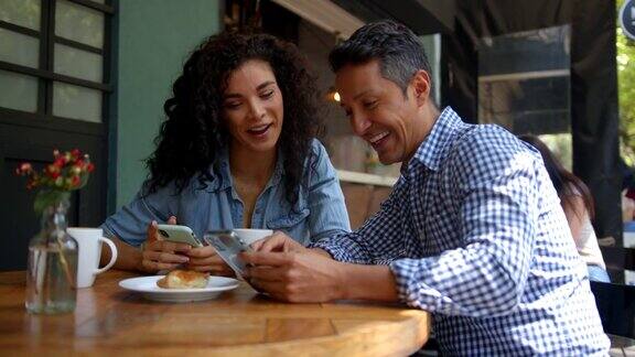 快乐的朋友们坐在咖啡店的户外桌子旁一边享受着咖啡和糕点一边看着他们的智能手机聊天和微笑