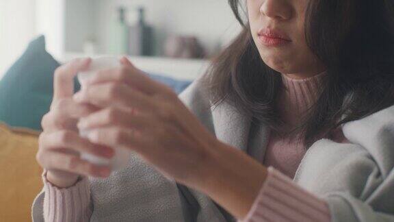 生病的亚洲妇女喝水喝处方药在家里客厅的沙发上盖着毯子忍受着头痛或抑郁近距离射杀