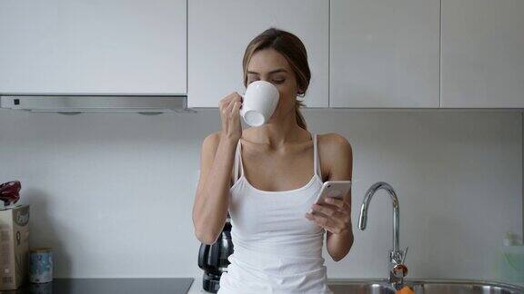 一名年轻女子靠在厨房柜台上一边喝着咖啡一边用智能手机发短信