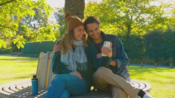 一对情侣坐在秋日公园的长椅上用手机进行视频通话