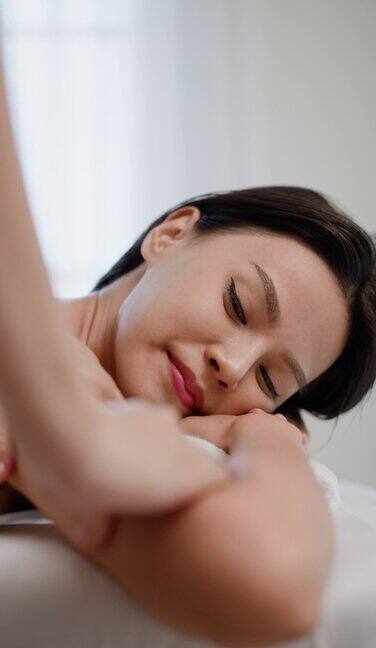 侧视图:年轻的亚洲女性与眼睛闭上躺在白色床水疗台接受手臂或身体按摩在健康水疗沙龙愉快的客户享受放松的背部理疗美容治疗