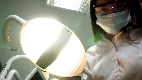 年轻女牙医戴着口罩带着工具走近牙科灯照进病人嘴里站在病人身上看着相机牙医的脸