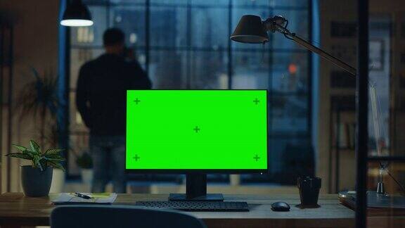 现代创意办公室的桌面电脑和绿色屏幕模型站在木桌上在背景中设计师从杯子里喝着饮料看着窗外的夜城