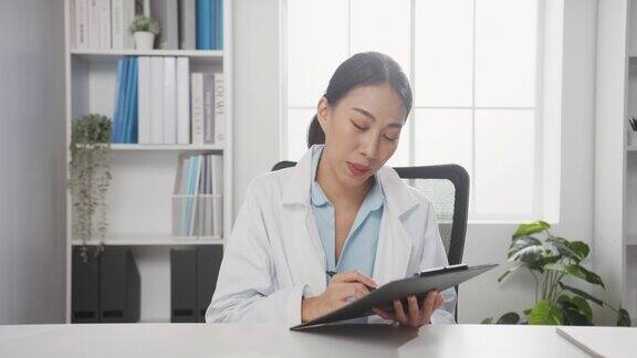 年轻的亚洲女医生穿着白色医疗制服带着听诊器用电脑笔记本电脑与病人进行视频会议看着摄像头在卫生院咨询和治疗理念