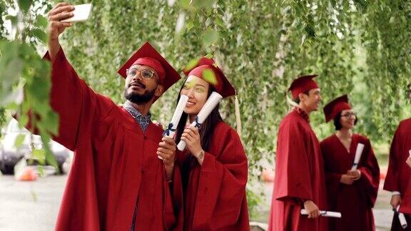 两名混血儿学生穿着学士帽和学士服拿着毕业证书自拍一名男子拿着智能手机拍照